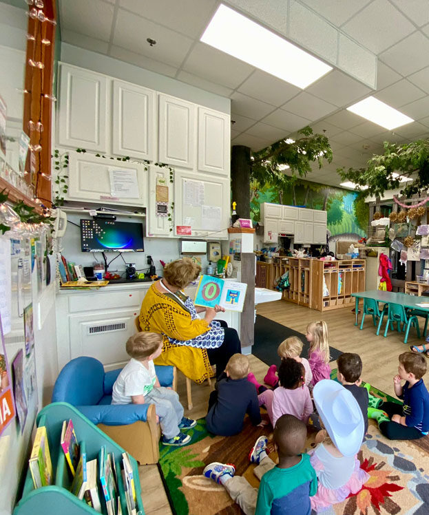 A picture of a teacher reading a book to 9 children sitting on the carpet below her in a thematically decorated Kids Junction classroom.