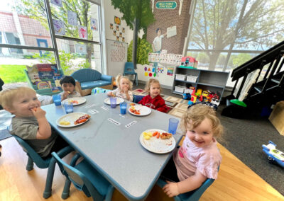 A picture of children in a classroom eating a meal at their table and smiling at the camera in a thematically decorated Kids Junction classroom.