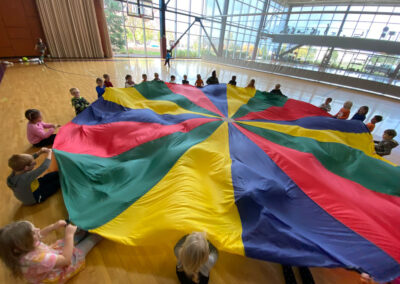 A picture of children sitting in a circle playing with a multi-colored parachute in the Princeton Club gymnasium.