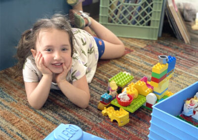 A picture of a little girl laying on her tummy on the floor of a classroom with her hands under her chin and playing with Legos.