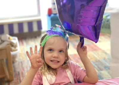 A picture of a little girl sitting at a table in a classroom with a bowl of treats in front of her, a birthday crown on her head and holding a birthday balloon while smiling at the camera.