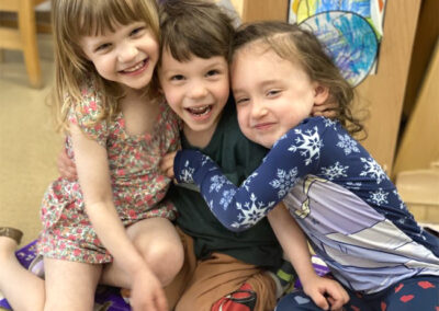 A picture of three little children on the floor of a classroom hugging and smiling at the camera.