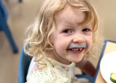 A little girl sitting at a table in a classroom with a plate and drink in front of her and smiling at the camera with a blue stain above her lip from the drink she is drinking.