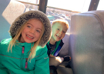 A picture of a little boy and a little girl sitting beside each other on the Kids Junction bus with their winter coats on and smiling wide at the camera.