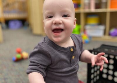 A picture of a little boy playing on the floor of an infant and toddler classroom and smiling at the camera.