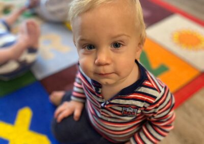 A picture of a little boy sitting on the floor of an infant and toddler classroom looking up at the camera and smiling slightly.