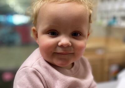 A picture of a little girl sitting on the floor of an infant and toddler classroom smiling at the camera.